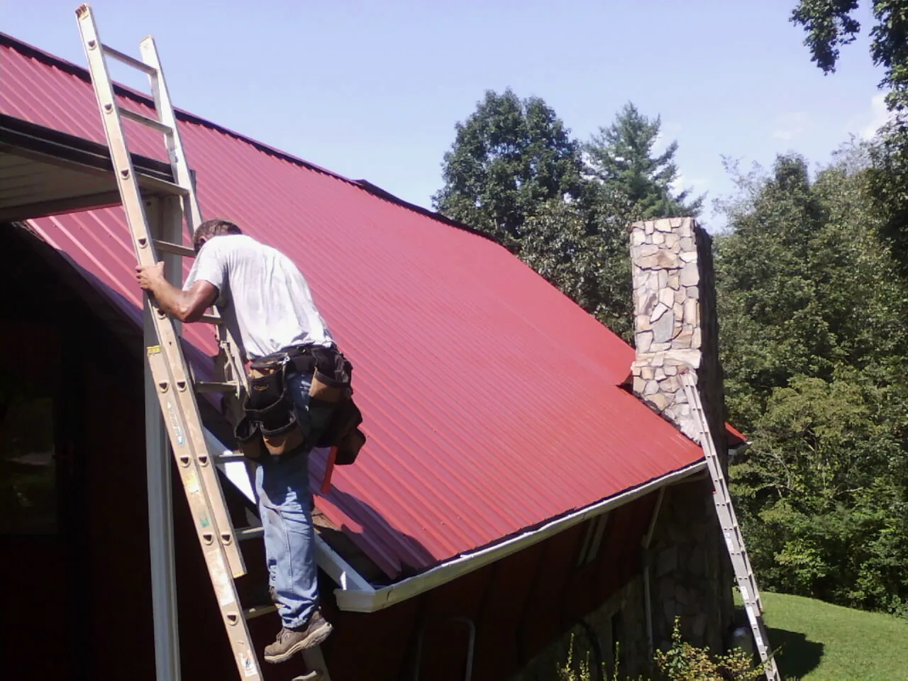 Roofer on a ladder beside a red metal roof and stone chimney — Soffit & Fascia Repair by Roofing Brothers in Nicholls